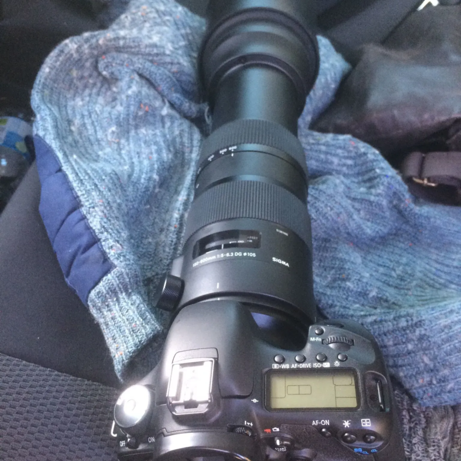 Macchina fotografica con obiettivo lungo su un sedile, accanto a un maglione.